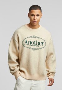 Beżowy sweter wykonany na drutach z okrągłym dekoltem, z zielonym logo "Another" i detalami wzorniczymi. Miękka tekstura, luźny krój.