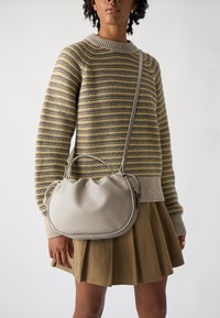 Beige skinnveske med rund design og rynket topp. Båret med en stripete genser og et brunt skjørt, som fremhever dens teksturerte overflate.