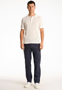 Vit kortärmad pikétröja med en strukturerad mönstring, tillsammans med mörkblå byxor och vita sneakers, stående mot en enfärgad bakgrund.