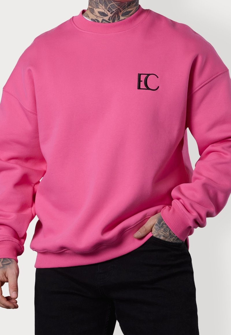 Personne portant un sweat-shirt ras du cou rose vif avec le logo noir "EC" sur la poitrine gauche et un pantalon noir, main dans la poche, tatouages visibles au cou et à la main.