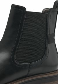 Botín Chelsea de cuero negro con panel lateral elástico texturizado, puntera redondeada y suela de madera marrón en contraste. Presenta una sutil marca en el elástico.