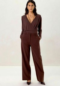 Brown wrap top with long sleeves paired with high-waisted, wide-leg brown trousers. Glossy pointed-toe heels complete the outfit.