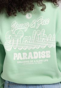 Mintgrüner Sweatshirt mit weißem Text: "Young & Free", "Malibu" und "Paradise". Der Text hat unterschiedliche Schriftarten und ein gestreiftes Muster.