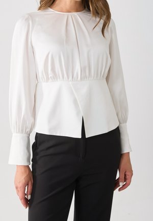 Frau trägt eine weiße Langarmbluse mit geraffter Taille und vorderem Schlitz, kombiniert mit schwarzer Hose vor einfarbigem Hintergrund.