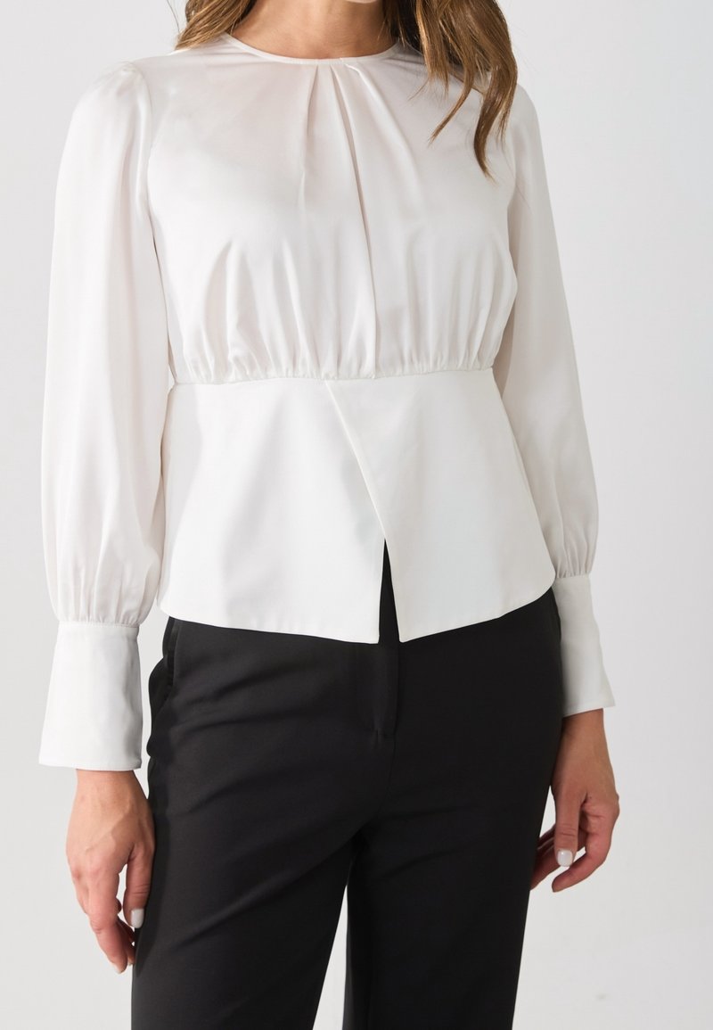 Femme portant un chemisier blanc à manches longues avec taille froncée et fente à l'avant, assorti d'un pantalon noir, sur un fond uni.