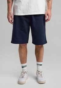 Tummansininen puuvillashortsit, joissa on sileä pinta, polvipituiset, viistetyt reunat. Yhdistetty valkoisiin tennareihin ja raidallisiin nilkkasukkiin.