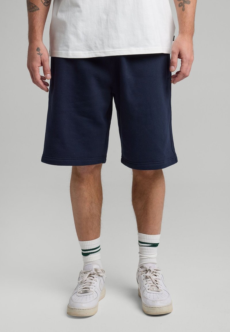 Tummansininen puuvillashortsit, joissa on sileä pinta, polvipituiset, viistetyt reunat. Yhdistetty valkoisiin tennareihin ja raidallisiin nilkkasukkiin.