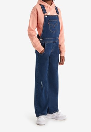 Barn iført blå denim overalls, ferskenfarvet hættetrøje og hvide sneakers, stående med den ene hånd i lommen mod en hvid baggrund.
