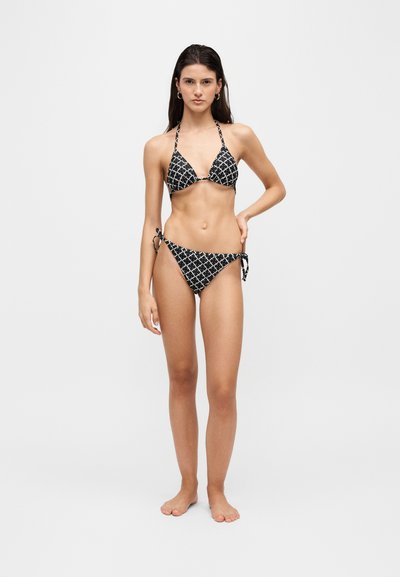 Jeune femme debout pieds nus portant un bikini string noir et blanc à motifs sur un fond clair uni.