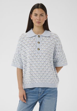 Donna con lunghi capelli scuri che indossa un maglione a maniche corte di colore azzurro chiaro con colletto e tre bottoni, abbinato a jeans blu.