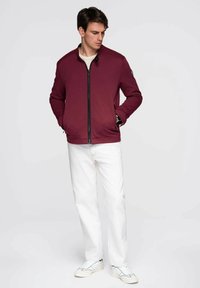 Burgunderfarbene Zip-Jacke aus strukturiertem Stoff, mit Stehkragen und schwarzen Reißverschlussdetails. Kombiniert mit hellen Jeans und weißen Sneakern.