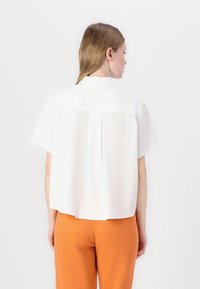 Camicetta bianca a maniche corte con colletto, dotata di una piega posteriore per una maggiore libertà di movimento. Abbinata a pantaloni arancioni a trama. Design minimalista.