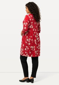 Rotes Blumenkleid mit Dreiviertelärmeln und tailliertem Oberteil, das an der Taille ausgestellt ist. Kombiniert mit schwarzen Hosen und Ballerinas. Glatte Stofftextur.