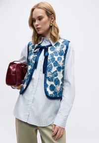 Νεαρή γυναίκα που φοράει λευκό ριγέ πουκάμισο και μπλε φλοράλ γιλέκο, κρατώντας μια μικρή κόκκινη τσάντα με χρυσούς κρίκους σκουλαρίκια.