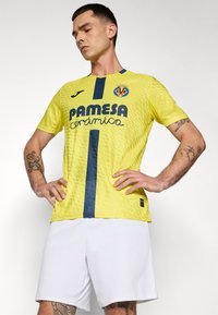 Mees atleet kannab kollast Villarreal CF-i särk, millel on tume sinine triip, ja valgeid lühikesi pükse, seismas käed puusas valge tausta ees.