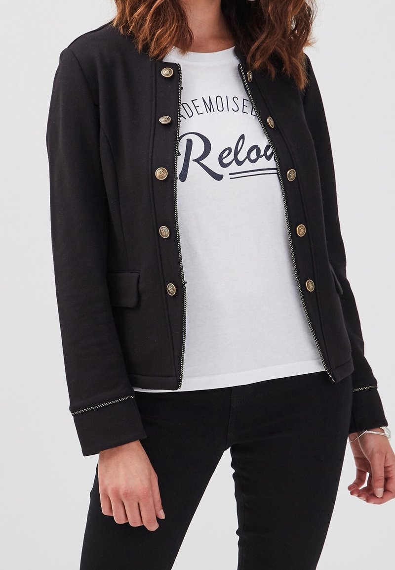 Black jacket with gold buttons, textured fabric, and a slim fit, layered over a white t-shirt featuring a printed design. Overall casual style.