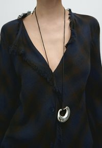 Marineblaues Shirt mit fransigen Kanten, Knopfdetails, offen getragen am Ausschnitt, mit einem silbernen Anhänger an einem schwarzen Band.
