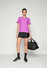 Lilla træningstrøje med korte ærmer, sammen med sorte shorts og sorte træningssko. En sort træningstaske bæres i højre hånd.