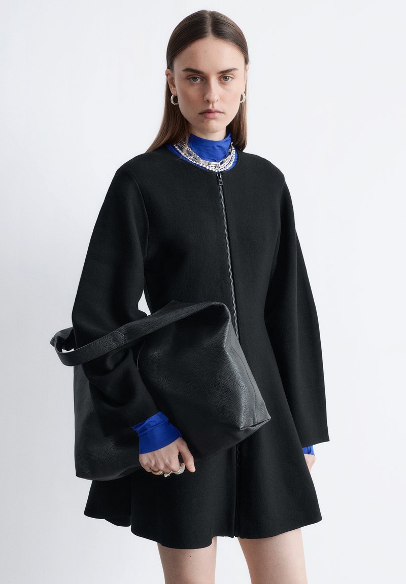 Manteau noir zippé avec des manches larges, porté sur un col roulé bleu. Le modèle tient un sac en cuir noir, avec une texture lisse et une sangle large.