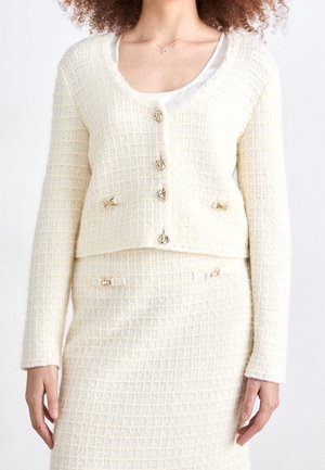 Mujer con cárdigan texturizado color crema con botones dorados y falda a juego con pequeños detalles de lazo dorado.