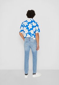 Korte mouwen blauwe shirt met wit bloemenpatroon, gecombineerd met lichtblauwe jeans en witte sneakers, bekeken vanaf de achterkant.