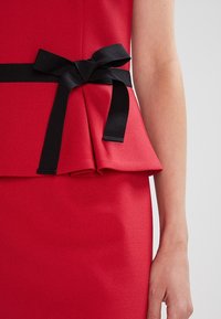 Haut peplum rouge avec un ruban noir noué à la taille. Le tissu est lisse, et le design comporte un détail peplum superposé à l'ourlet.