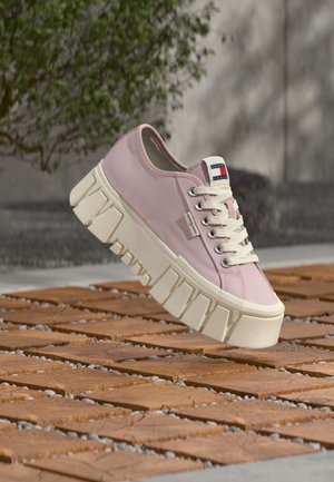 Lys rosa lavtoppet joggesko med tykke hvite klumpete såler og hvite snøringer, vist utendørs på en overflate av tre og småstein.