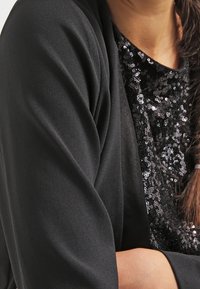 Kobieta w czarnej bluzce z cekinami i czarnym żakiecie, z warkoczem opadającym na ramię, z ręką złożoną na ciele.