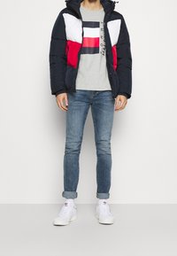 Chaqueta tipo puff en azul marino, blanco y rojo; diseño acolchado con cierre de cremallera y capucha. Camiseta gris con gráfico; vaqueros azules y zapatillas blancas.