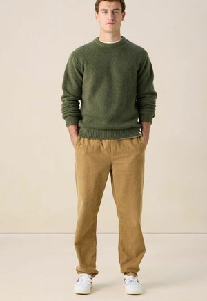 Man staat met handen in zijn zakken, gekleed in een groene trui, tankleurige corduroy broek en witte sneakers, voor een effen beige achtergrond.