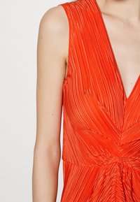Rotes, plissiertes Kleid mit tiefem V-Ausschnitt, das vertikale Linien aufweist, die sich an der Taille vereinen und eine strukturierte, fließende Erscheinung erzeugen.