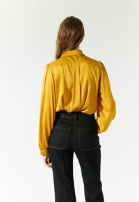 Blusa di raso gialla con maniche a sbuffo, schiena plissettata e colletto, abbinata a jeans in denim neri a vita alta con cuciture visibili.