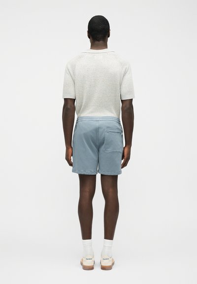 Homme portant une chemise en maille beige clair à manches courtes, un short bleu avec une poche arrière, des chaussettes blanches et des baskets beiges, debout de dos.
