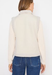 Gilet en polaire crème avec des accents gris, un col et un intérieur doux. Design sans manches, l'ourlet arrive à la taille, associé à un jean en denim bleu.