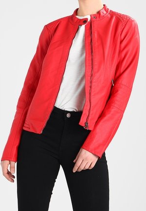 Kobieta ubrana w czerwoną skórzaną kurtkę na białą koszulę i czarne spodnie, stojąca na tle jednolitego jasnego tła.