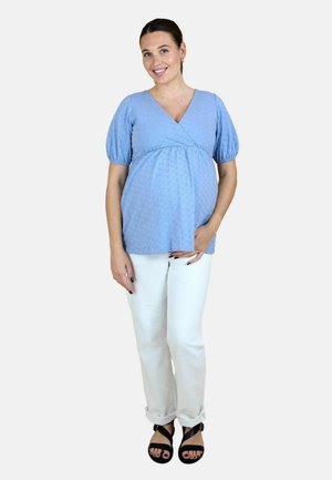 Zwangere vrouw die een lichtblauw blouse met korte mouwen en een witte broek draagt, glimlacht met één hand op haar buik en zwarte sandalen.