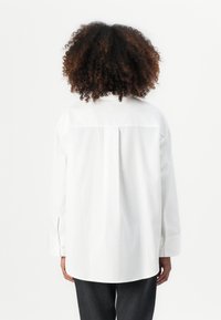 Camisa blanca de manga larga con un corte relajado, que presenta una costura central en la parte posterior y puños abotonados. Textura de tela suave, sin patrones.