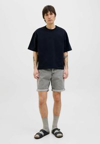 T-shirt noire à manches courtes associé à un short en denim gris, avec des poignets retroussés, des chaussettes grises jusqu'aux genoux et des sandales noires à lanières.