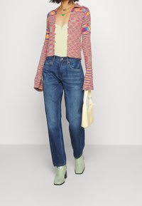 Cárdigan de punto multicolor con mangas acampanadas, bodysuit con cuello en forma de scallop, jeans rectos azules, botas verdes hasta el tobillo y un bolso amarillo.