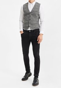 Grå houndstooth väst med ett subtilt mönster, fyra mörka knappar, bärs över en vit skjorta med krage, i kombination med svarta slim-fit jeans och svarta skor.