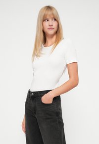 Jeune femme aux longs cheveux blonds portant un haut ajusté à manches courtes blanc et un jean noir taille haute, regardant sur le côté avec une main dans la poche.