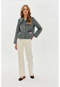 Šedá kostkovaná bunda s volánkovým límcem, zlatými knoflíky a dvěma kapsami s růžovou nášivkou, nošená s krémovými kalhotami a tmavými botami.