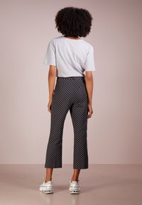 Kvinna med lockigt hår som bär en ljusgrå t-shirt, högmidjade svarta mönstrade byxor med cropped längd och vita sneakers, stående mot en enfärgad bakgrund.