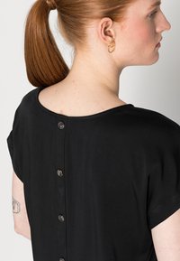 Blusa preta com mangas curtas, decote arredondado e uma linha de botões nas costas. Tecido suave com um brilho sutil.