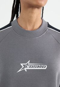 Osoba oblečená v šedém svetru s kulatým výstřihem, na ramenech má černobílé pruhy a bílé hvězdné logo s textem "COSMO".