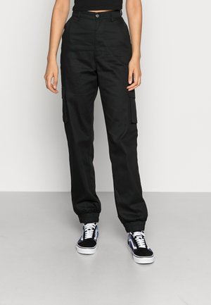 Femme portant un pantalon cargo noir taille haute avec des poignets élastiques et des baskets noires et blanches à lacets, debout sur un sol gris.