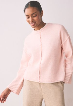 Femme aux cheveux foncés en chignon bas, portant un pull rose doux et un pantalon beige, souriante et regardant vers le bas dans une pose détendue.
