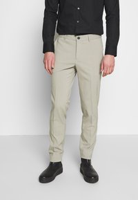Pantaloni beige sartoriali con vestibilità dritta, dotati di chiusura con bottone verde, tasche laterali e un tessuto liscio e leggero.