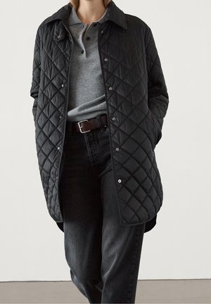 Person wearing a black quilted jacket over a gray buttoned sweater, black belt, and dark washed black jeans, standing with hands in pockets.