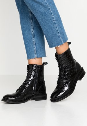 Glänzende schwarze Schnür-Stiefeletten mit Krokodilprägung und seitlichen Reißverschlüssen, getragen zu Jeans mit ausgefransten Säumen auf hellem grauem Boden.